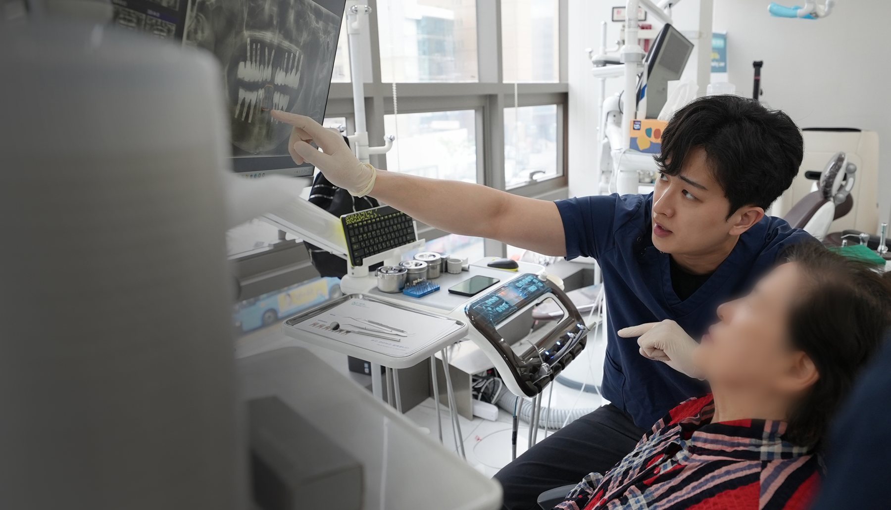 남색 진료복 의사가 X-ray 모니터를 가리키며 환자에게 설명하는 장면(환자 얼굴 흐림)